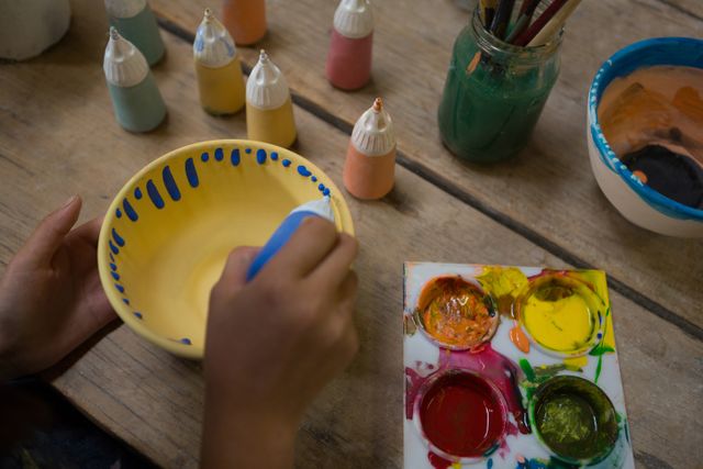 Child painting a ceramic bowl in a pottery workshop. Various paint bottles, brushes, and a palette with vibrant colors are on the wooden table. Ideal for use in articles about children's activities, art and craft workshops, creative hobbies, and educational programs.