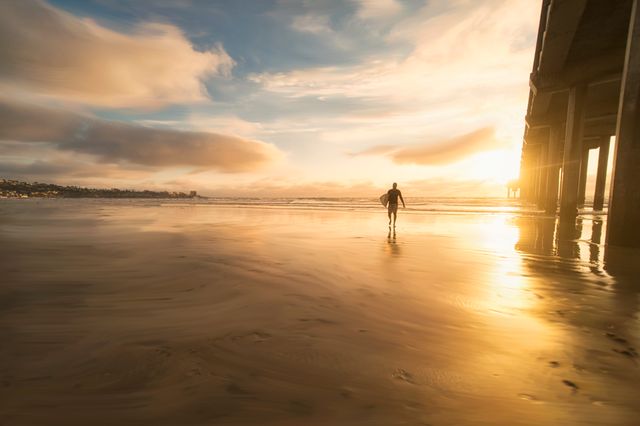 Man walking along sandy beach during sunset near pier, creating silhouette with golden hues reflecting on wet sand. Ideal for travel promotions, beach lifestyle ads, inspirational content, and nature photography showcases.