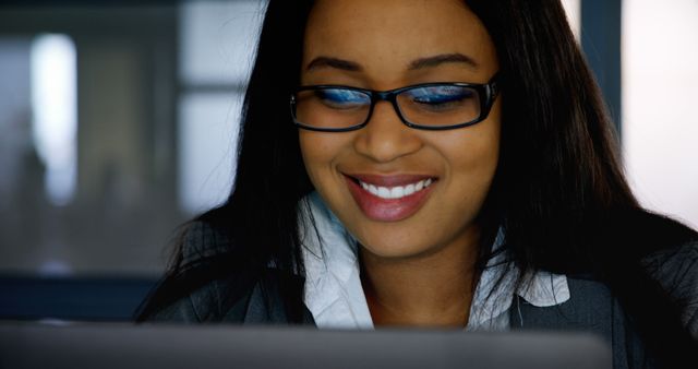 African American businesswoman smiling while working on a laptop in a modern office environment. Useful for illustrating professional, corporate, and business-related themes such as success, productivity, and workplace happiness.