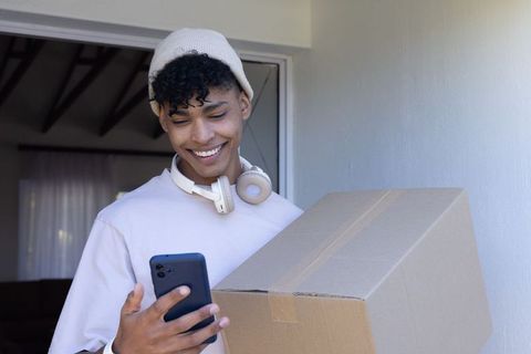 African american man carrying parcel checking smartphone at home doorway, smiling