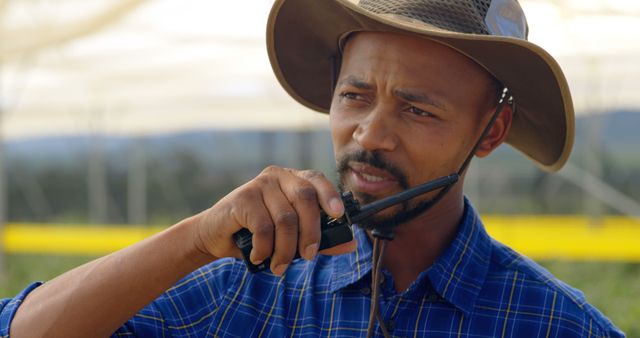 Man wearing a wide-brimmed hat and blue plaid shirt is communicating using a walkie talkie in an outdoor environment. Could be useful for topics related to outdoor activities, agriculture, field work, rural settings, or communication technology.