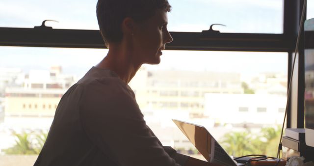 Woman using tablet next to window in modern office with cityscape view. Perfect for business, technology, or corporate lifestyle themes. Useful for illustrating concepts of remote work, digital workspaces, or modern workplaces.