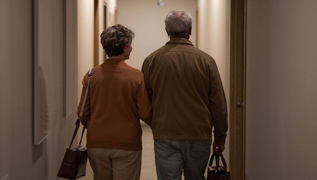 Senior couple walking arm-in-arm in apartment corridor carrying handbags wearing warm sweaters