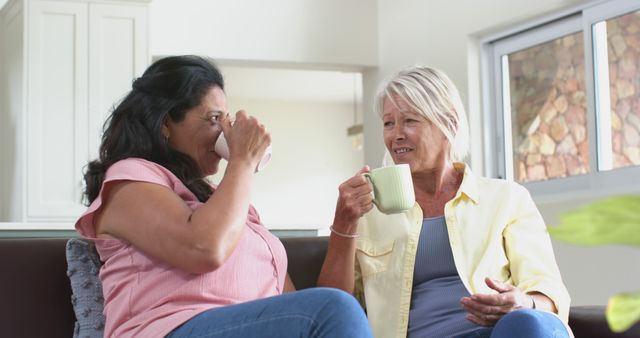 Two mature women are sitting in a living room, smiling and enjoying coffee together. They are casually dressed and appear relaxed. Great for use in articles or advertisements related to friends bonding, casual home life, relaxed conversations, and products aimed at mature adults.