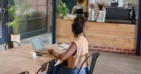Woman raising hand while working on laptop in bright coffeehouse with papers and espresso
