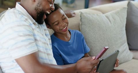 African american father and son laughing, sharing smartphone and tablet on couch