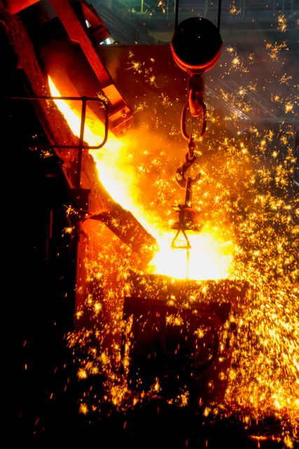 Molten metal pouring from a furnace inside an industrial foundry with bright sparks flying. This is an ideal depiction of heavy industry, metalworking, and high heat environments. The intense energy and dynamic motion make it suitable for use in marketing materials for manufacturing companies, educational displays about metallurgy, or illustrating articles related to industrial processes or engineering feats.
