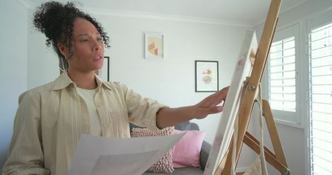 African woman painting on easel at home studio holding sketch and adjusting canvas