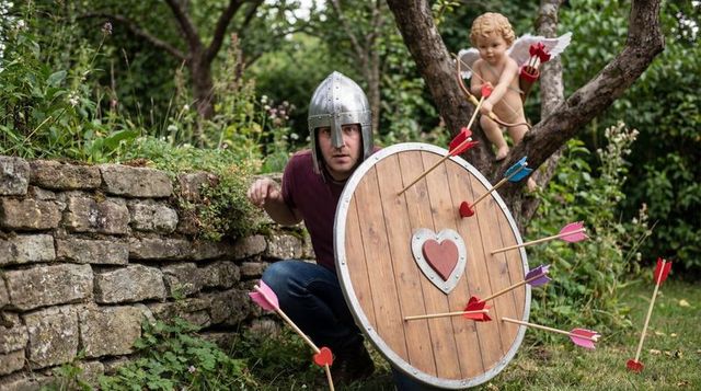 Crouching man wearing medieval helmet hiding behind heart shield while cupid fires arrows