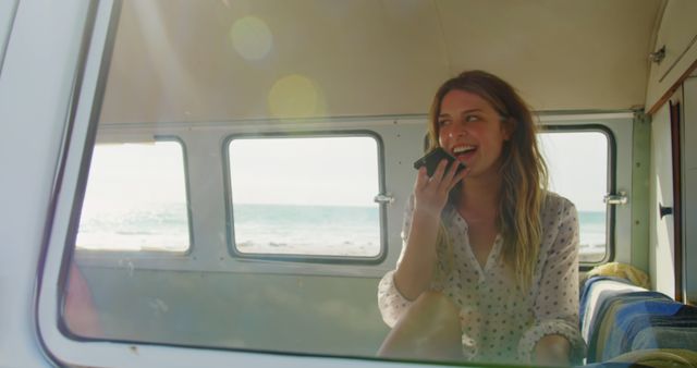 Young woman sits inside a camper van parked by the ocean, talking on the phone and smiling. The background shows an ocean view through the van's windows, with sunlight flooding into the van. Perfect for illustrating themes of adventure, travel, freedom, joy, and a relaxed lifestyle. Suitable for use in travel blogs, advertisements, lifestyle articles, and social media content promoting outdoor and beach activities.