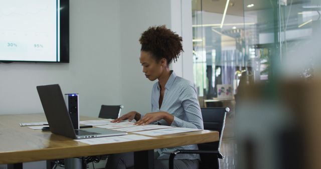 Biracial businesswoman sitting at desk, checking documents and using laptop in office. business professional and working in busy modern office.