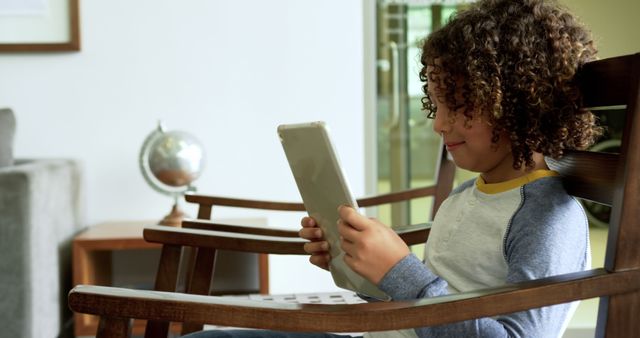 Young boy with curly hair sitting on a wooden chair in a modern living room, engaged with a tablet. Suitable for themes related to education, technology, children's activities, home lifestyle, and digital learning.