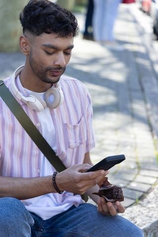 Young indian man sitting on curb checking smartphone with headphones and snack