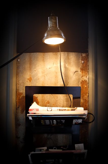 This image features a warmly lit, cozy reading nook with an industrial-style lamp casting a soft glow on a collection of books and magazines neatly arranged on metal shelving. Perfect for articles about creating comfortable reading spaces at home, interior design inspirations, or decorating small spaces with an industrial touch.
