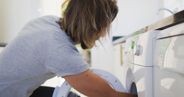 This image shows a young person loading laundry into a washing machine in a domestic setting. The casual clothing suggests a relaxed, everyday activity. Ideal for articles, advertisements, or instructional materials related to home care, appliances, daily routines, or sustainable living.