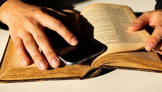Hands Resting on Open Aged Book with Smartphone Nearby Showing Digital vs Analog Contrast