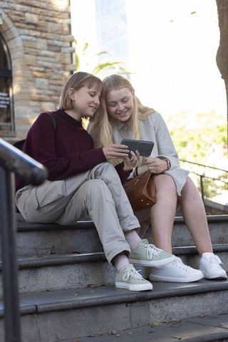 Two young women sharing smartphone on urban stone steps laughing and chatting