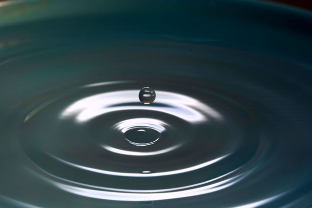 This image captures the precise moment when a water drop splashes into a calm water surface, creating ripples emanating outward. The focus on the droplet and ripples highlights themes of tranquility, purity, and the beauty of small natural events. Suitable for use in environmental campaigns, wellness and skincare products, photography art, and educational materials discussing fluid dynamics or nature's beauty.