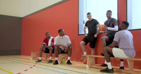 Multiracial basketball teammates talking and resting on bench in indoor gym