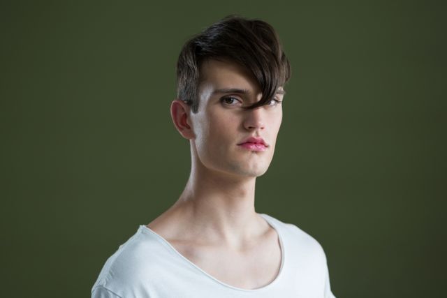 This image features an androgynous man with a modern hairstyle, looking directly at the camera against a green background. His serious expression and stylish appearance make this image suitable for use in fashion magazines, contemporary lifestyle blogs, or advertisements focusing on modern androgynous beauty standards. The minimalistic background and white shirt add to the clean and sophisticated look, making it versatile for various editorial and commercial purposes.