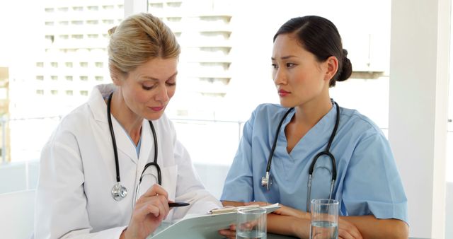 An older female doctor in a white coat with a stethoscope is mentoring a younger female nurse in blue scrubs. They are indoors in a modern healthcare facility, with the doctor attentively explaining details on a clipboard and the nurse listening intently. This imagery is ideal for illustrating medical education, mentoring, teamwork, and training in the healthcare industry. It can be used in healthcare training programs, hospital advertisements, medical articles, and educational materials.