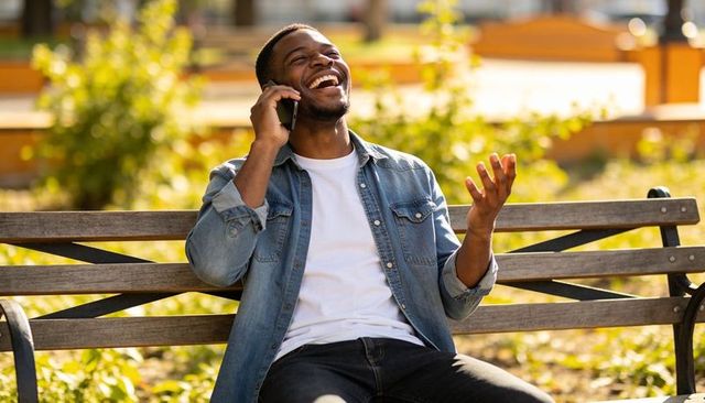 Laughing young man talking on smartphone sitting on wooden bench in sunny urban park plaza