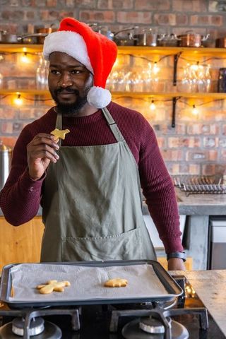 African american man wearing santa hat and apron holding cookie while baking in cozy brick kitchen