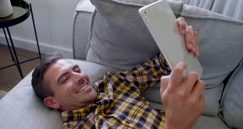 Man Happily Gazing at Tablet While Relaxing on Sofa