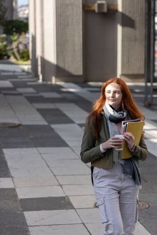 Redhead woman walking urban plaza holding coffee and notebooks in sunlit shadowed walkway
