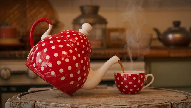 Pouring red polka-dot enamel teapot into matching cup on rustic wooden table