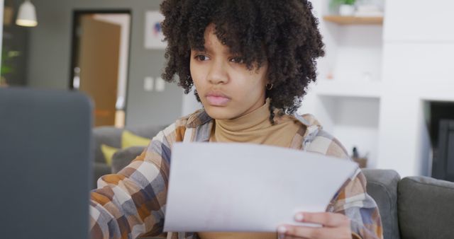 A young woman with curly hair is concentrating while working on a laptop, holding a piece of paper. She is dressed casually in a plaid shirt and is in a home office environment. This image is great for topics related to remote work, study, online learning, and productivity. It could be used for articles or advertisements about remote working conditions, managing work from home, or effective study practices.