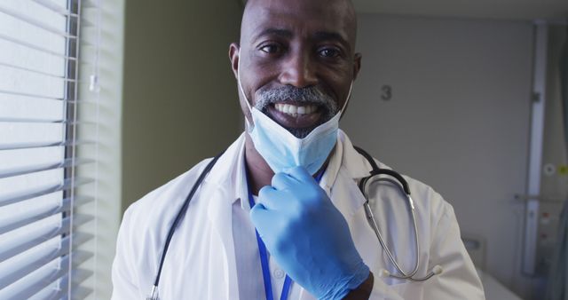 Portrait of smiling african american male doctor wearing face mask standing in hospital room. medicine, health and healthcare services during coronavirus covid 19 pandemic.