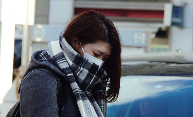 Woman wearing a protective mask and plaid scarf on city street, offering clues into preventive health measures. Suitable for themes around winter clothing, urban lifestyle, public safety, health awareness, and casual fashion in winter. It can be used for articles, blog posts, or health and safety campaigns.