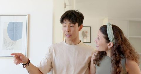 Asian man pointing at framed art while african american woman admiring in sunlit modern home