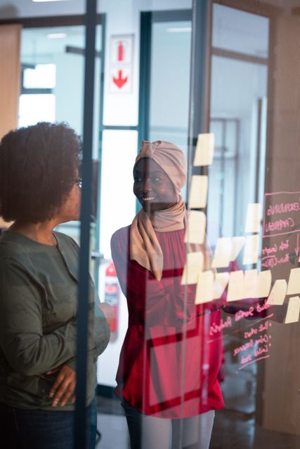 Two happy african american female colleagues brainstorming by notes on glass wall in office. working in business at a modern office.