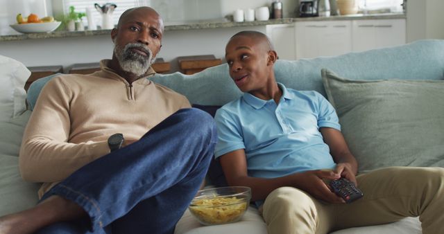 Father and son sitting on a sofa in a living room, watching TV together, and smiling. They are both relaxed and casually dressed. This image can be used to depict family, bonding, quality time, and home life. Useful for ads, blog posts, or digital content centered on family relationships, leisure activities, or home living.