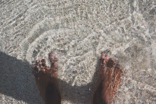 This image shows a close-up of two bare feet standing in clear shallow water with ripples forming around them. Ideal for use in content related to relaxation, beach vacations, mindfulness, and connecting with nature. It can be used to represent peaceful moments, outdoor activities, and the simplicity of enjoying nature’s elements.