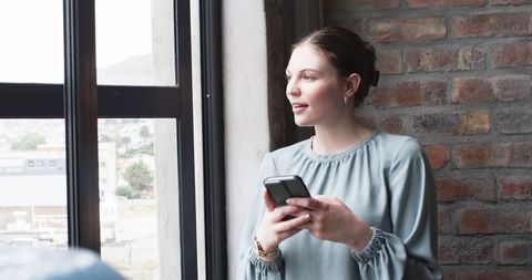 Young Professional Using Smartphone Near Window in Relaxed Office Setting - Download Free Stock Images Pikwizard.com