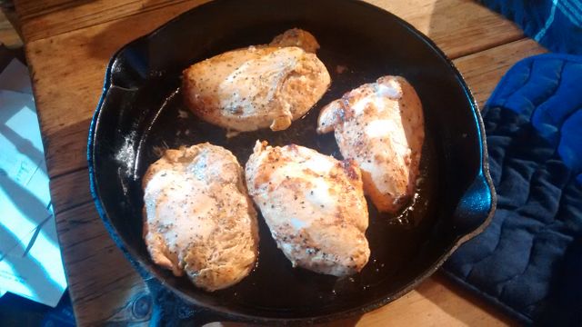 Four seasoned chicken breasts sizzling in cast iron skillet on wooden table. Ideal for illustrating home-cooked meals, healthy eating, and rustic kitchen settings. Perfect for recipe blogs, cooking websites, and meal preparation tutorials.