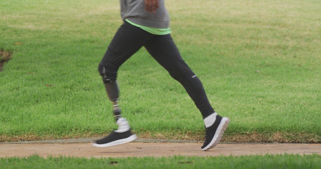 Individual wearing a prosthetic leg jogging on a path in a green park. Ideal for promoting fitness, rehabilitation, healthy lifestyle, and outdoor exercise activities.