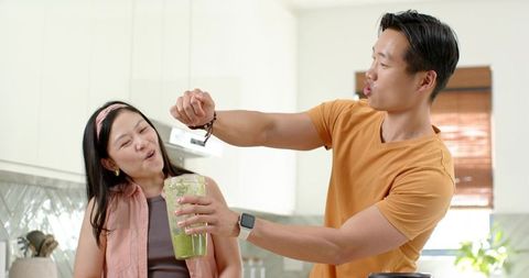 Asian couple preparing green smoothie in modern kitchen, man sprinkling spice playfully