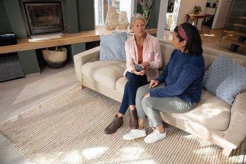 Multicultural mature women chatting on cozy living room sofa while sharing smartphone
