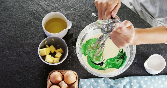 Hands seen from above mixing green food coloring into batter using whisk. Ingredients like butter, eggs, cooking oil, and milk container arranged around. Ideal for illustrating baking concepts, recipes, cooking blogs, or DIY projects.