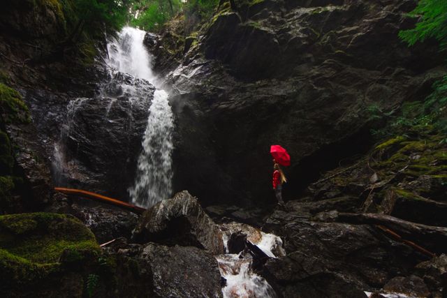 Person standing near a powerful waterfall surrounded by lush green forest, holding a bright red umbrella. Mist from waterfall creates a dramatic atmosphere, showcasing the beauty of nature and solitude. Perfect for themes related to adventure, exploration, tranquility, outdoor activities, and inspiring journeys.