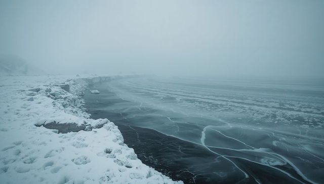 Snow-Covered Rocky Shoreline Curving into Fog Over Frozen Lake with Cracked Ice Sheets