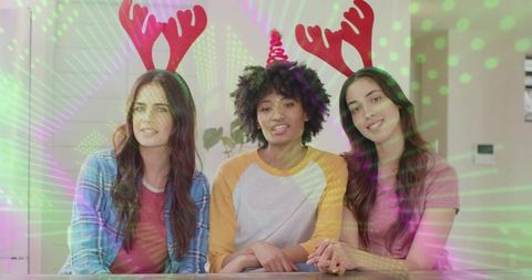 Laughing friends wearing reindeer antlers and party hat at kitchen table under colorful laser lights