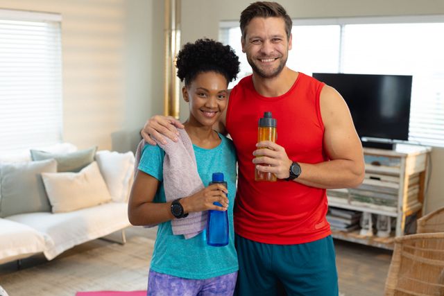Diverse couple smiling and holding water bottles while exercising in a living room. Ideal for promoting home fitness routines, healthy lifestyle choices, and quality time spent together. Suitable for use in fitness blogs, wellness websites, and advertisements for home workout equipment.