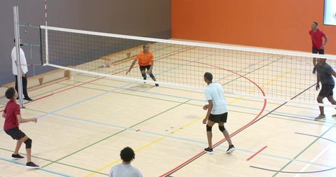 Diverse men playing indoor volleyball on light wood court wearing knee pads