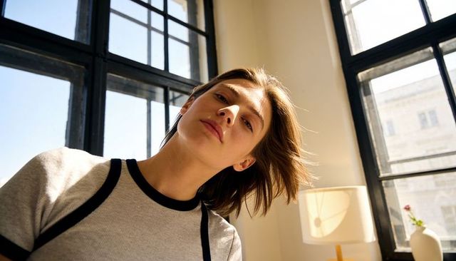 Leaning woman tilting toward camera in sunlit modern urban apartment with grid windows