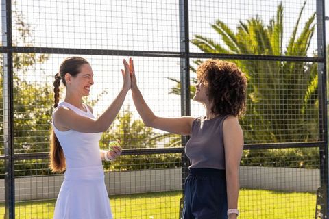 Female tennis partners high-fiving courtside holding ball outdoors sunny fitness smiling